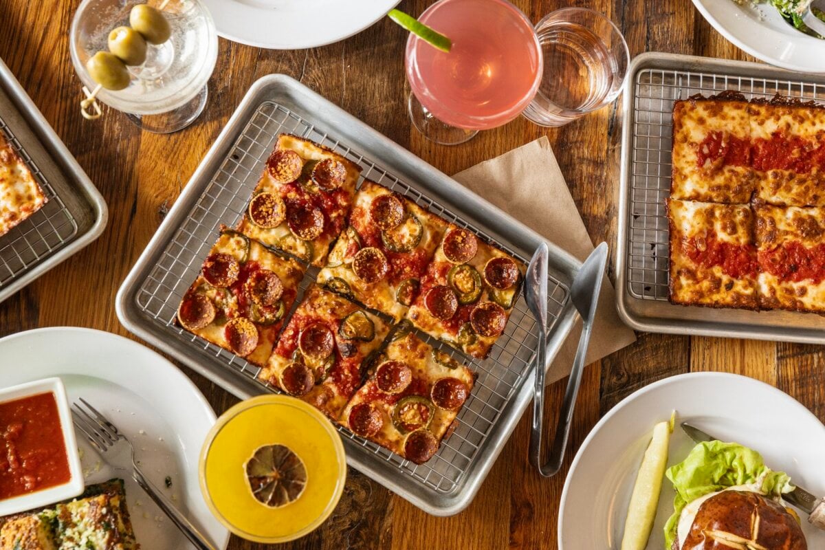 3 New Louisville Restaurants to Try this Month (+2 New Menus!) - 6 Rectangular pizzas with pepperoni and tomato sauce are served on trays, surrounded by cocktails, a salad, and dipping sauce on a wooden table.