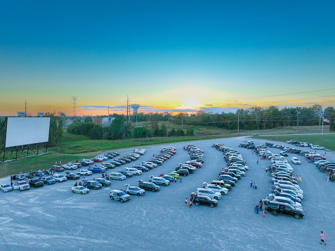 5 Louisville Summer Movie Series Not to Miss - 5 Rows of cars are parked at a Louisville drive-in theater at sunset, with a large movie screen and people walking near vehicles, capturing the classic charm of summer outdoor movies.