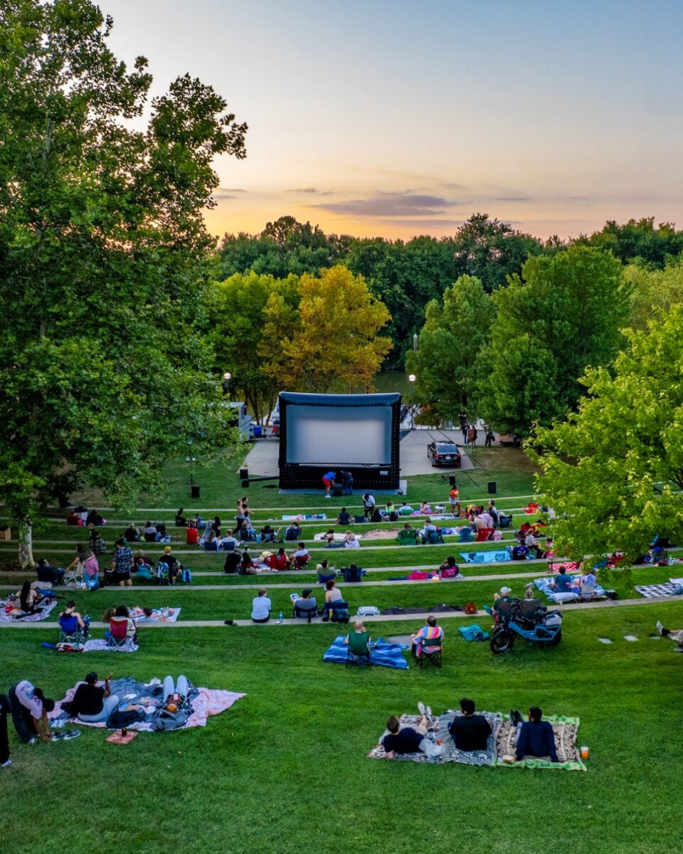 5 Louisville Summer Movie Series Not to Miss - 6 People sit on blankets and lawn chairs in a park, enjoying outdoor movies on a large inflatable screen at sunset during a classic Louisville summer evening.