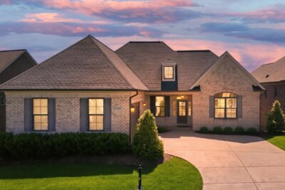 Single-story brick house at 15019 Tradition Drive, Louisville, KY, features multiple gables, glowing windows, a manicured lawn, and a curved driveway beautifully illuminated at sunset.