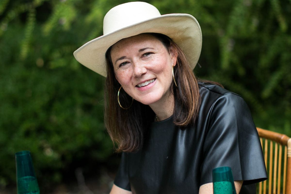 Cassie Lazzo sits outdoors at a table in a white hat and black top, smiling warmly at the camera with lush greenery behind her.