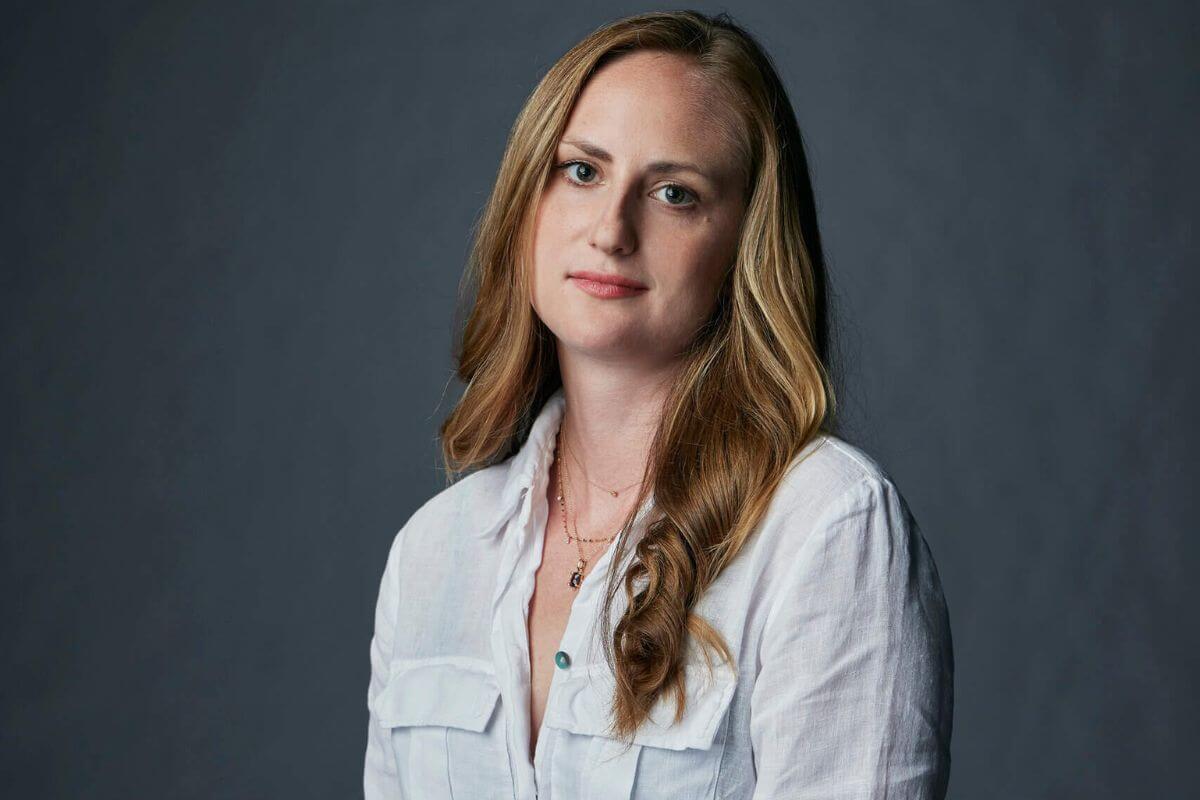 Suzannah Herbert, a woman with long, wavy blonde hair wearing a white button-up shirt, poses against a plain gray background.