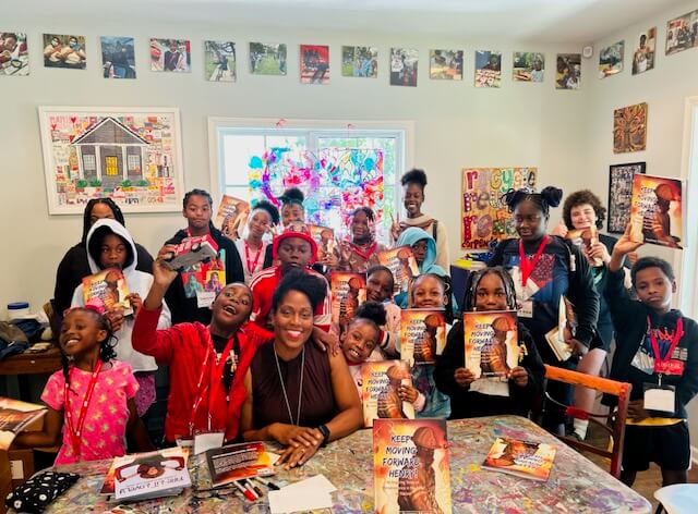 She Shapes Creativity & Community at Carpenter Art Garden - 6 A group of children and adults smile and hold up copies of Ayanna Murray's book "Keep Moving Forward" in a colorful, art-filled room.
