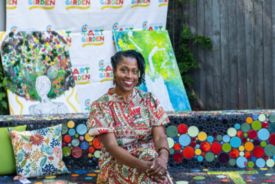 A woman in a patterned dress sits on a colorful mosaic bench at the "Community Art Garden," with abstract paintings and a banner by Ayanna Murray in the background.