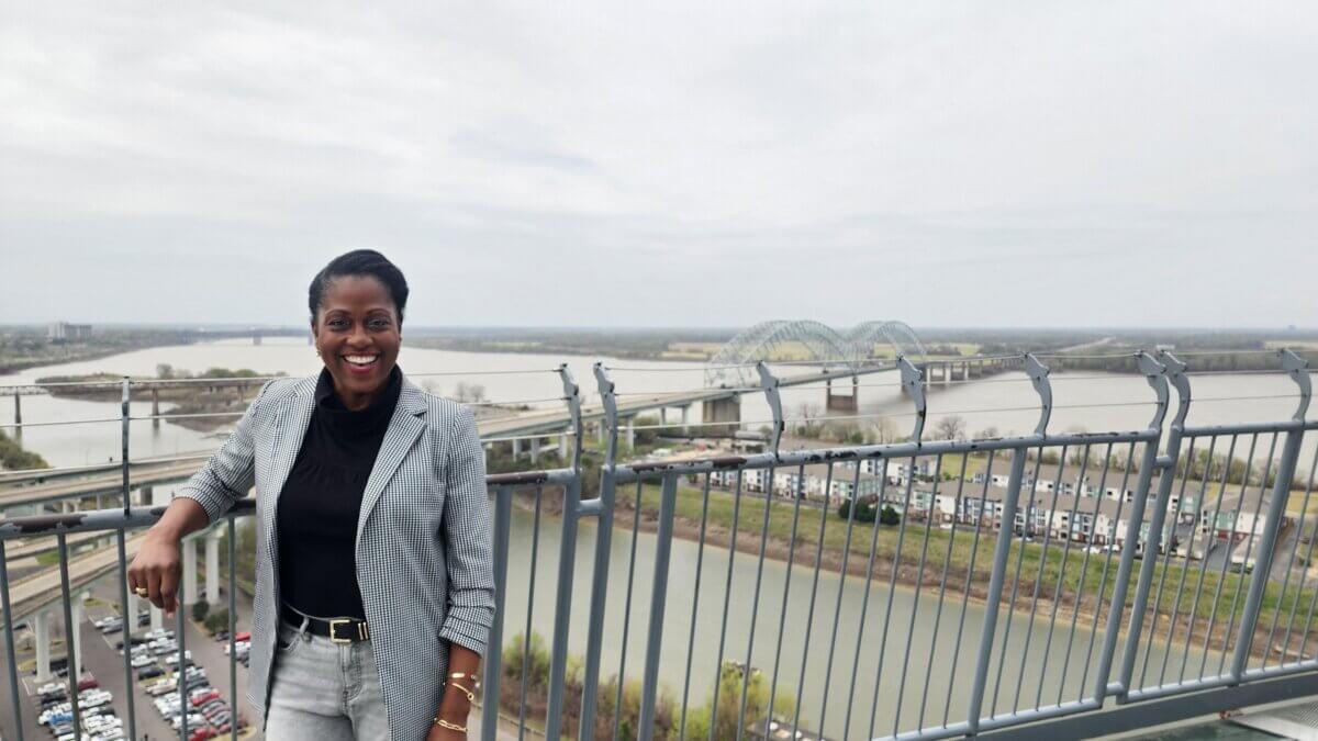 She Shapes Creativity & Community at Carpenter Art Garden - 7 Ayanna Murray stands smiling on a high observation deck overlooking a river, a bridge, and a parking lot with cloudy skies in the background.