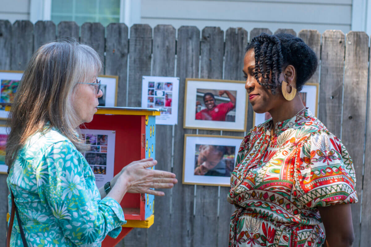 She Shapes Creativity & Community at Carpenter Art Garden - 4 Two women stand and talk in front of a wooden fence displaying framed photos at an outdoor event.