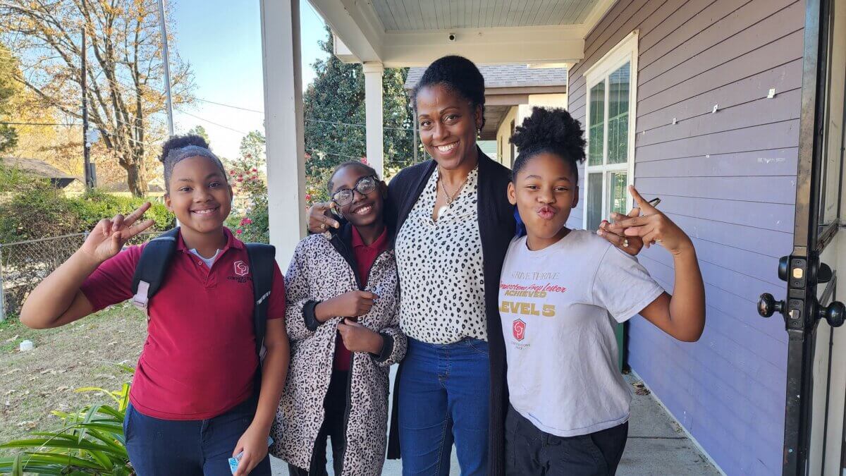 She Shapes Creativity & Community at Carpenter Art Garden - 3 Four people stand on a porch smiling at the camera; two children make peace signs with their hands. Trees and a purple house wall are visible in the background.