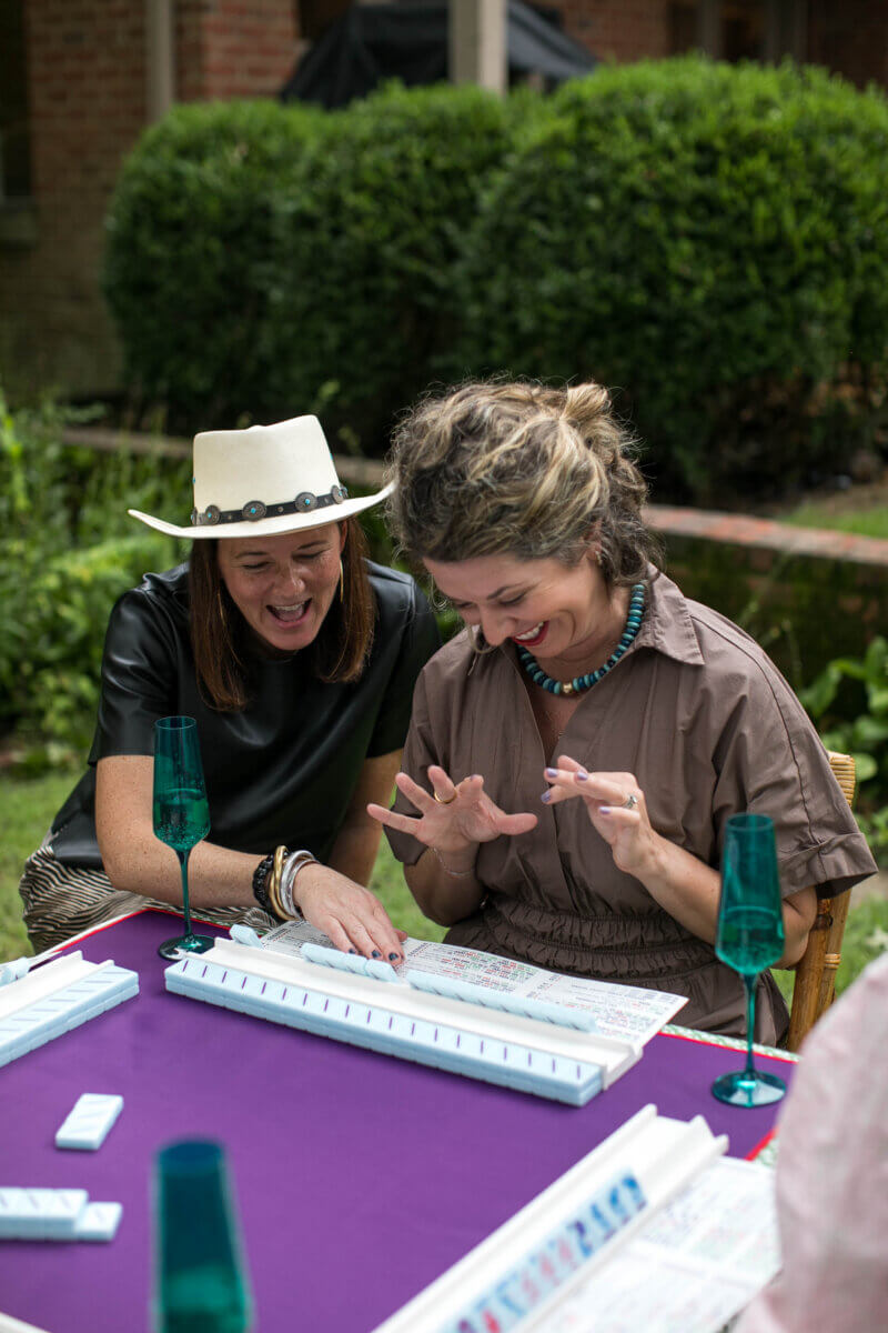 Meet "The Mahjologist" of Memphis, Cassie Lazzo - 3 Cassie Lazzo and a friend sit at a table outdoors, smiling as they look at game tiles in front of them, with teal glasses and a purple tablecloth.