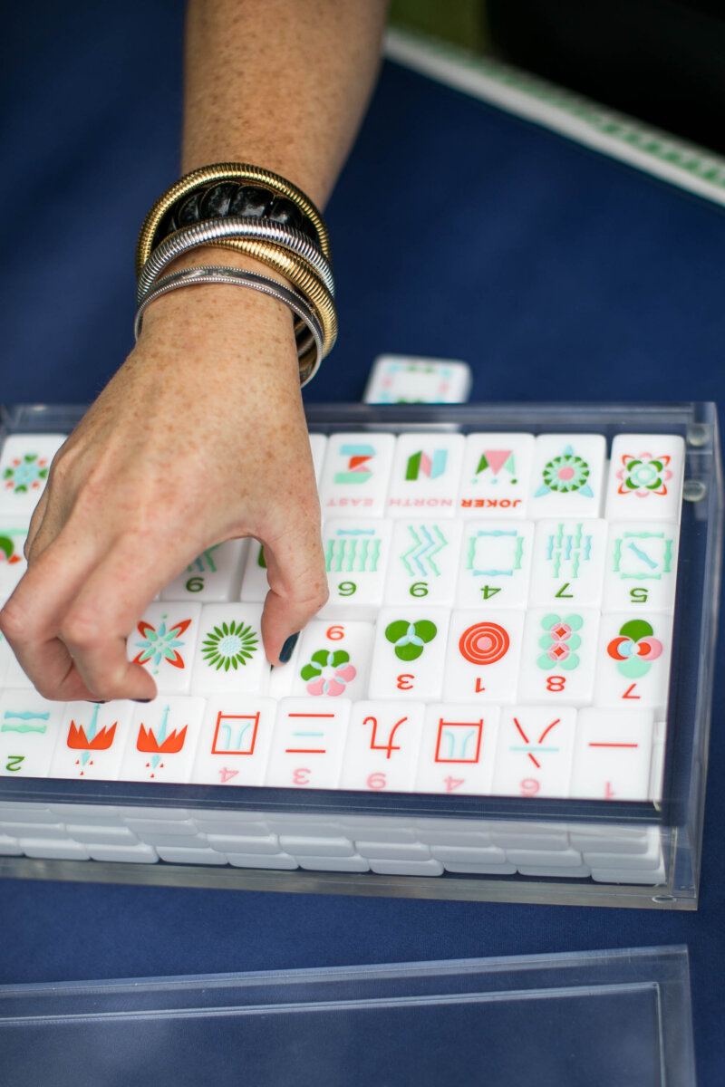 Meet "The Mahjologist" of Memphis, Cassie Lazzo - 4 A hand wearing bangles is picking up a Mahjong tile from a tray of arranged tiles on a blue surface.