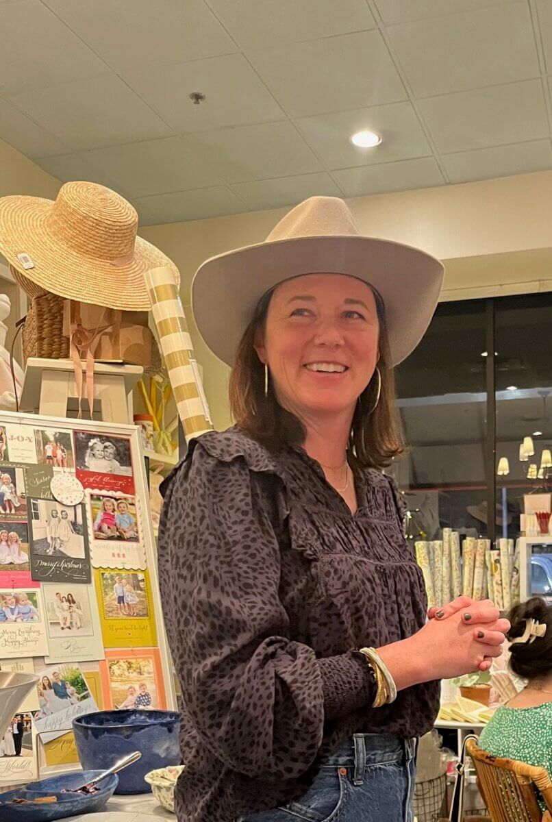 Meet "The Mahjologist" of Memphis, Cassie Lazzo - 2 A woman wearing a gray hat and patterned blouse stands indoors, smiling, with greeting cards and various merchandise displayed in the background.