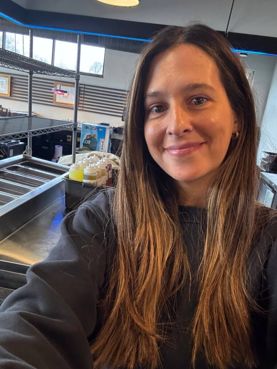 How Kelcie Nollner Built a Beloved Memphis Salad Destination - 2 A woman with long brown hair wearing a dark sweatshirt takes a selfie in a commercial kitchen or food prep area with stainless steel counters and supplies in the background.
