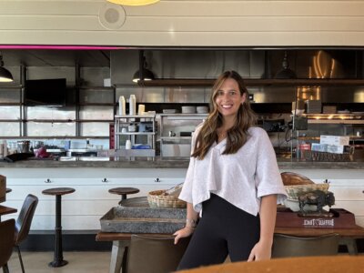 Kelcie Nollner stands with her long brown hair cascading down as she poses in front of a counter in a modern, well-lit restaurant with industrial-style decor.