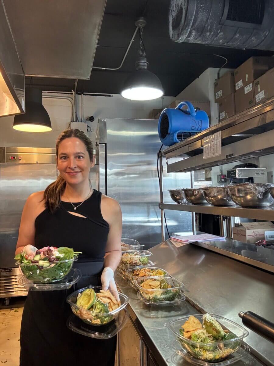 How Kelcie Nollner Built a Beloved Memphis Salad Destination - 3 A woman wearing a black top and gloves holds a salad bowl in a commercial kitchen, with several prepared salads lined up on the counter beside her.