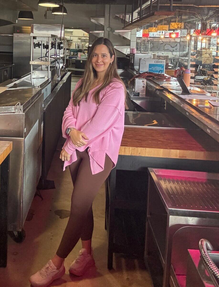 How Kelcie Nollner Built a Beloved Memphis Salad Destination - 5 A woman with long brown hair, wearing a pink sweater, brown leggings, and pink sneakers, stands in an empty restaurant kitchen near a counter.