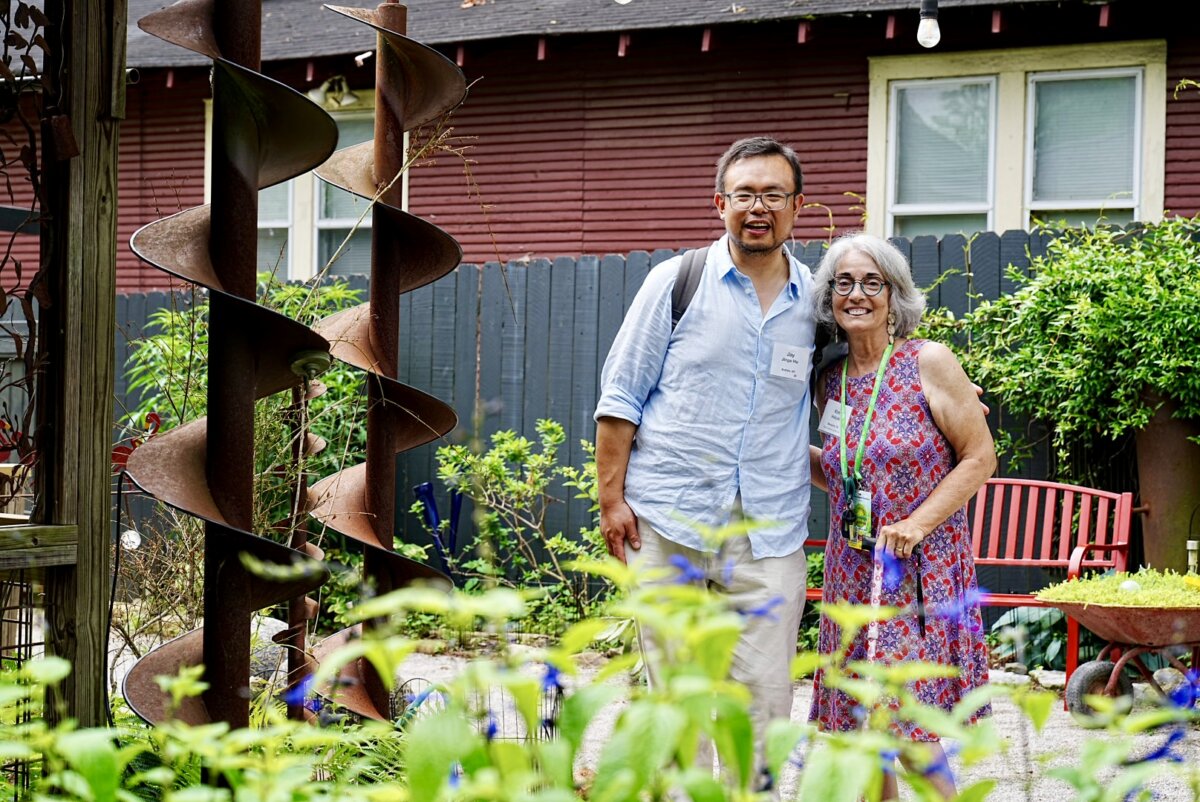 Two people stand together and smile in a lush garden, surrounded by plants, a large metal sculpture, and a red wooden bench. Created by Kim Halyak for FACES, the scene features a house and fence in the background.