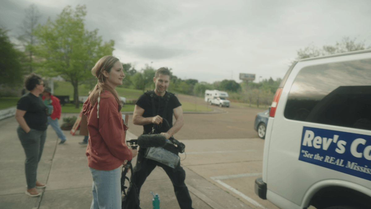 This Memphis Filmmaker is Reframing the Southern Story - 3 A woman stands next to a man holding audio equipment near a van labeled "Rev's Cafe" in a parking lot, with other people and greenery in the background.