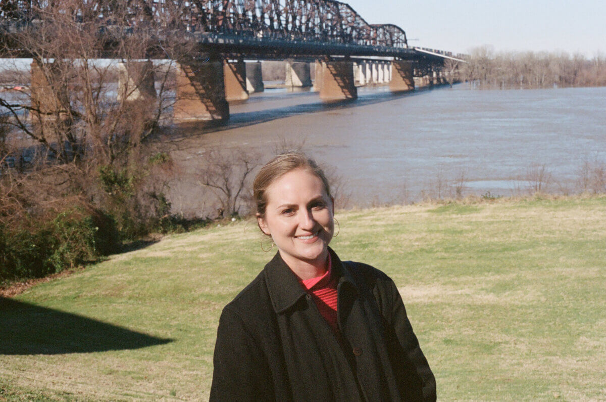 This Memphis Filmmaker is Reframing the Southern Story - 2 A woman in a black coat stands on a grassy area near a river, with a large steel bridge and trees in the background under a clear sky.