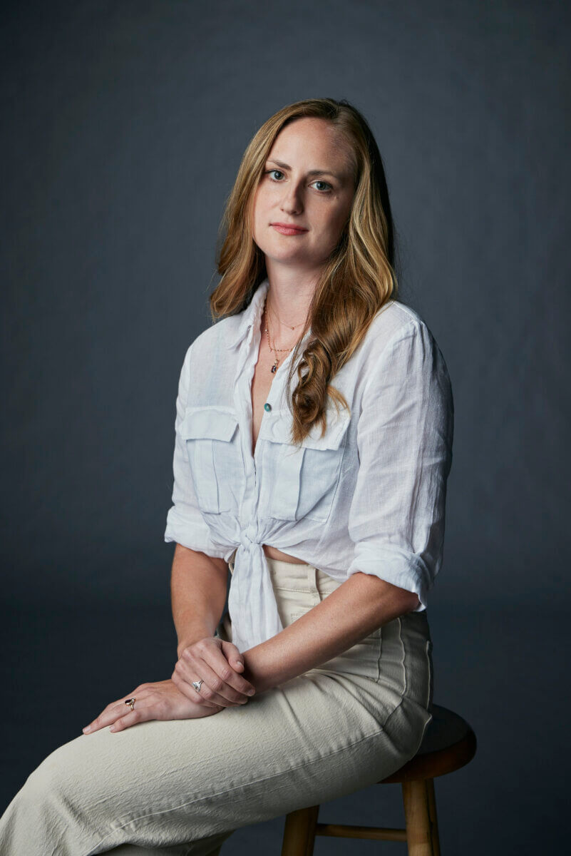 This Memphis Filmmaker is Reframing the Southern Story - 7 Suzannah Herbert, with long, wavy blonde hair, wearing a white button-up shirt and light pants, sits gracefully on a stool against a plain dark background.