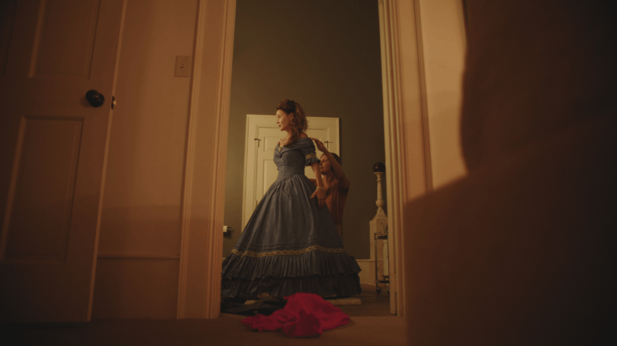 This Memphis Filmmaker is Reframing the Southern Story - 6 A woman in a blue, old-fashioned dress stands in a room; her coat draped over her shoulder. In the foreground, a pink garment lies on the floor.