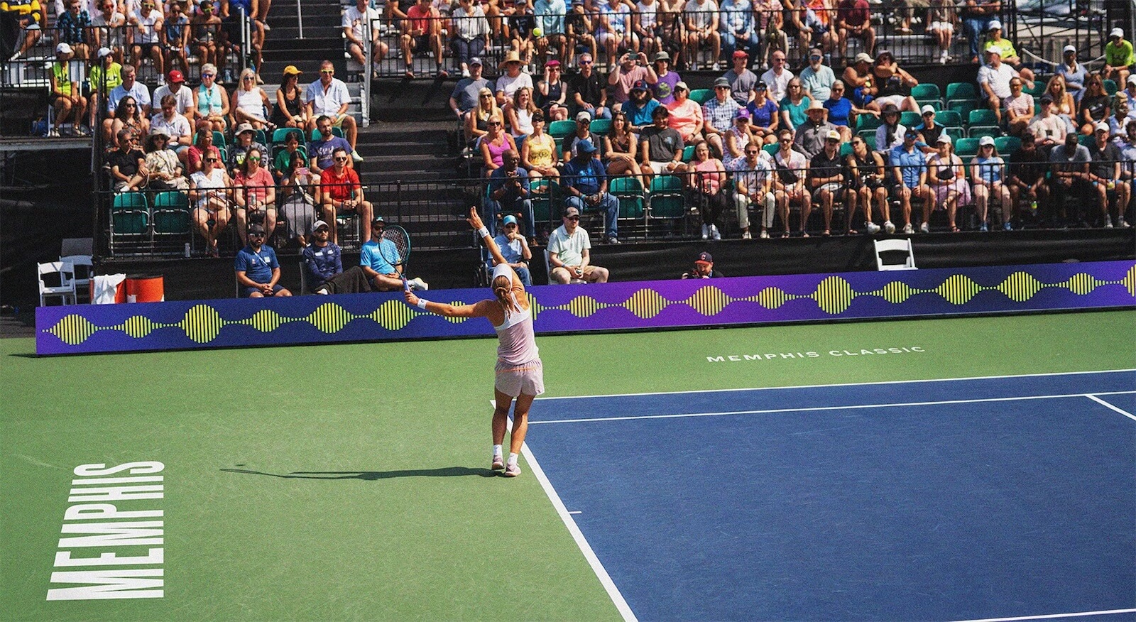 A tennis player serves on an outdoor court labeled “Memphis” during the Memphis Classic, with a large audience watching from the stands behind in Bluff City.