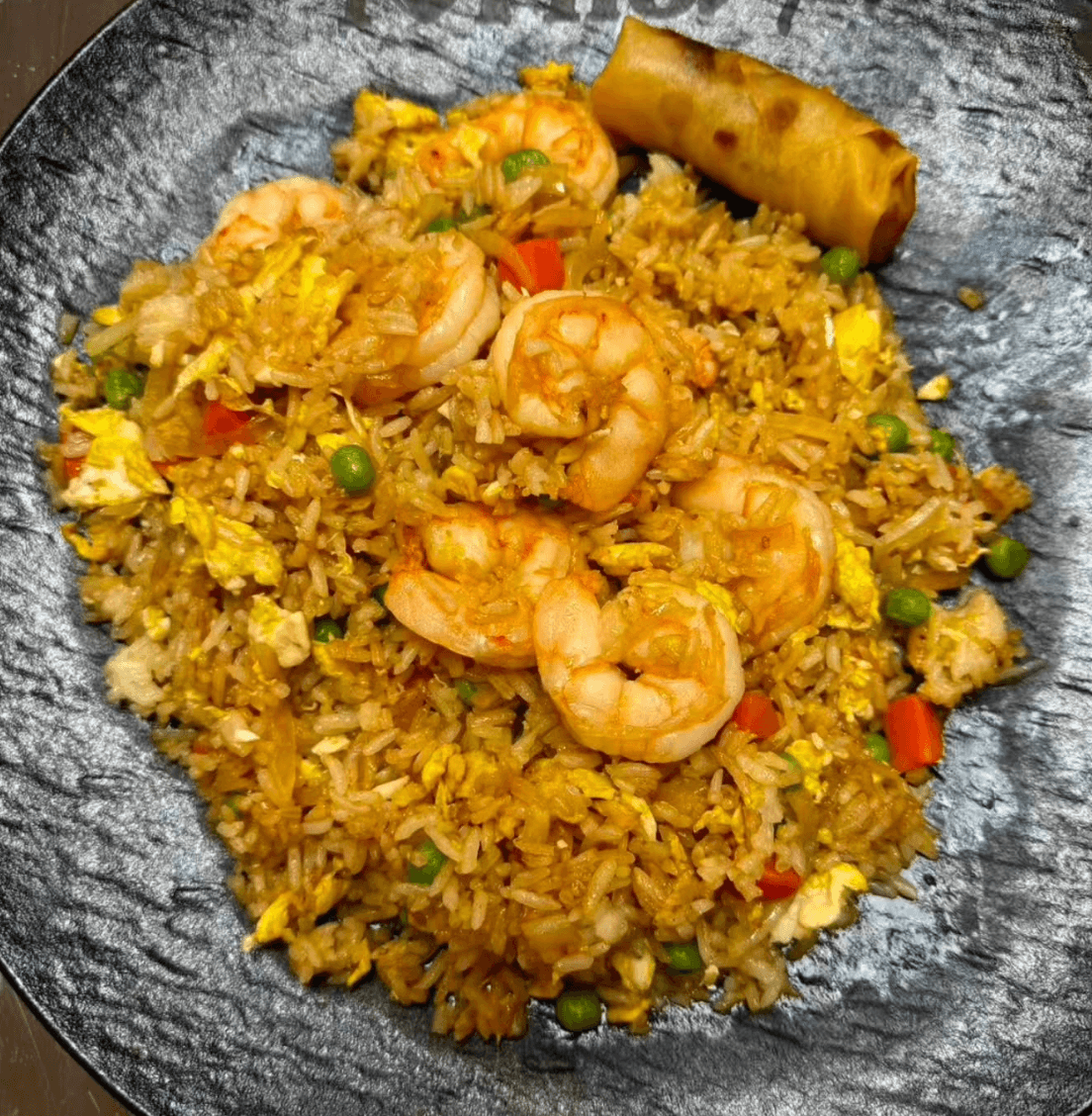 A plate of shrimp fried rice with vegetables and scrambled eggs, topped with several shrimp, served with a single fried spring roll.
