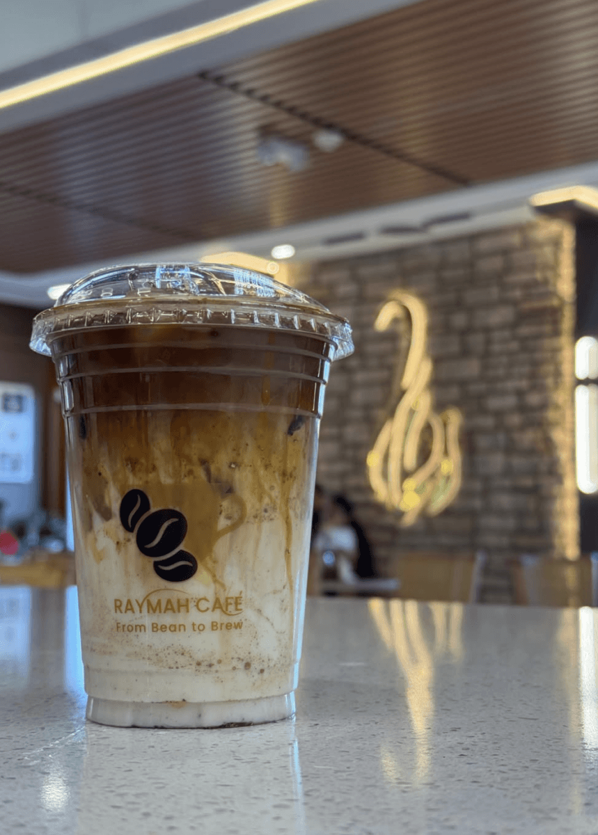 A plastic cup of iced coffee with visible milk and espresso layers sits on a counter inside Raymah Café, with a blurred café interior in the background.