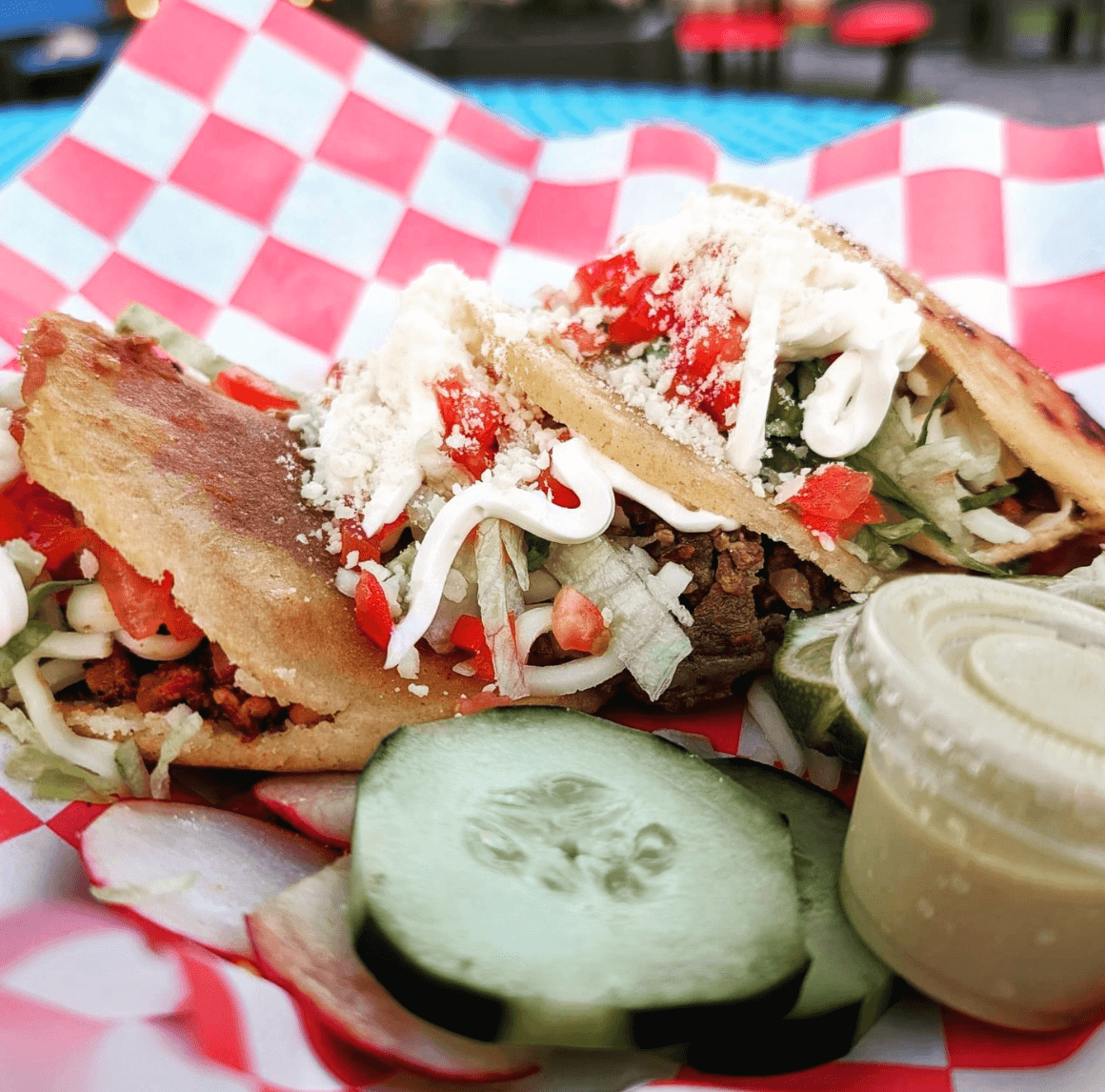 Two stuffed tacos topped with cheese, sour cream, and diced vegetables are served on a checkered tray with cucumber slices and a small cup of green sauce.