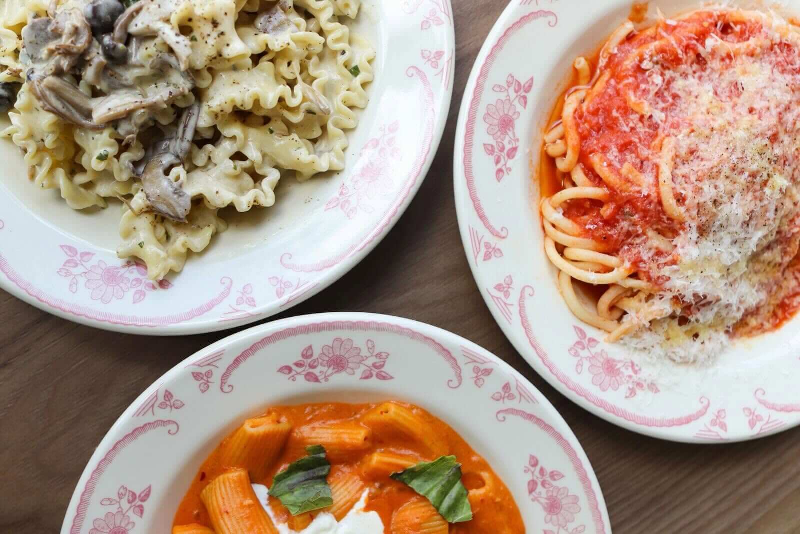 Three plates of pasta: one with creamy mushroom sauce, one with tomato sauce and grated cheese, and one with rigatoni in orange tomato sauce with basil.