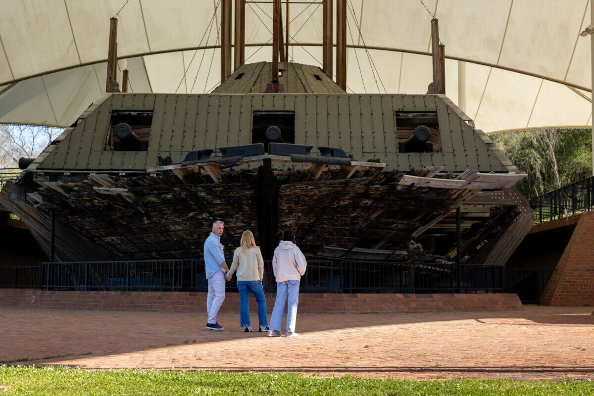 Three people stand in front of the preserved remains of a historic ironclad warship displayed under a large canopy.