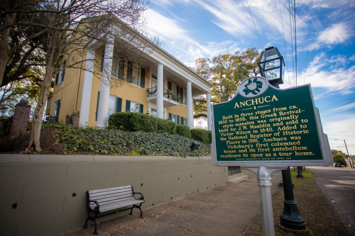 A historic yellow antebellum house with white columns stands behind a green and yellow Anhcuca information sign and a bench on a sidewalk in Vicksburg, Mississippi.