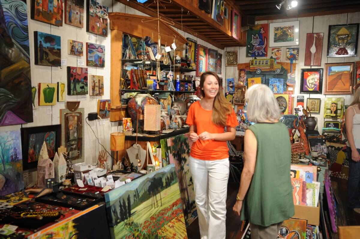 Two women talk inside a colorful art gallery in Vicksburg, Mississippi, surrounded by paintings, sculptures, and various handmade items displayed on walls and tables.