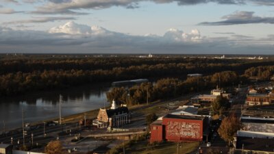 A river runs alongside the Mississippi town of Vicksburg, with trees in the background, buildings in the foreground, and a red mural that reads "Welcome to Vicksburg" on one building.