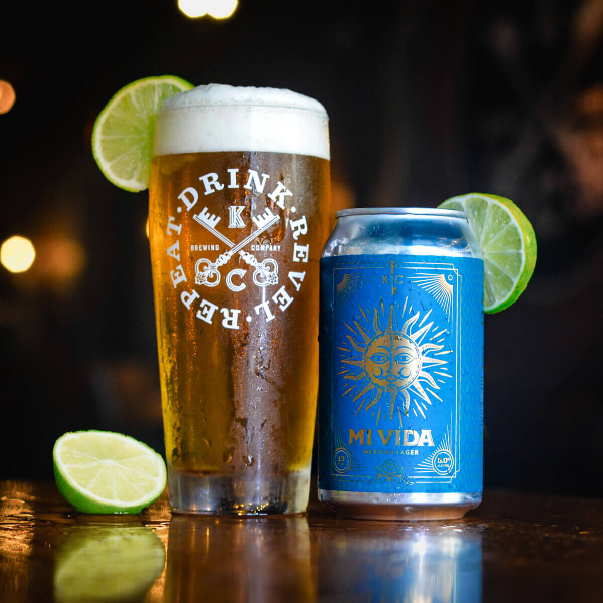 A glass of lager with a foamy head and lime wedge sits next to a blue "Mi Vida" can on a wooden surface, capturing the laid-back vibe of Vicksburg, Mississippi, with blurred lights in the background.