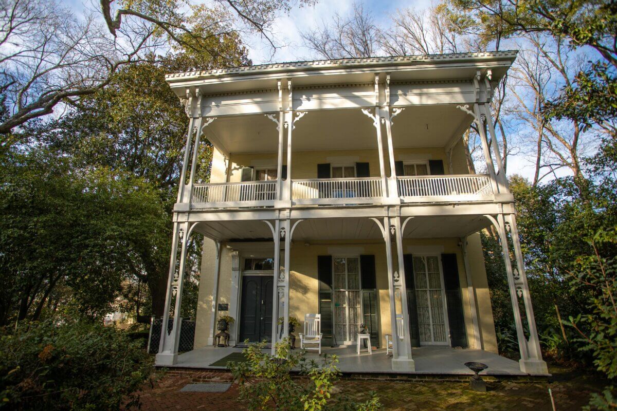 Two-story historic house in Vicksburg, Mississippi, featuring a double front porch with white columns and railings, surrounded by trees and lush greenery.