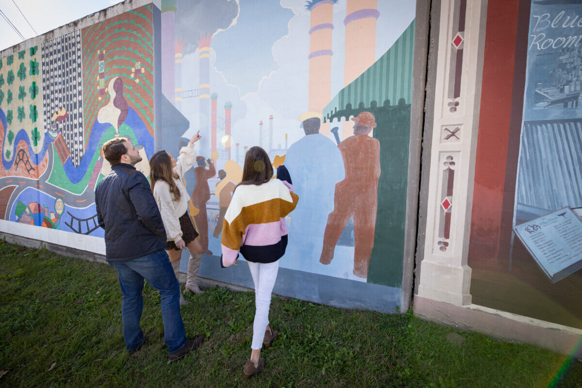 Three people stand on grass in Vicksburg, Mississippi, looking at a colorful mural on an outdoor wall, with one person pointing at the artwork.