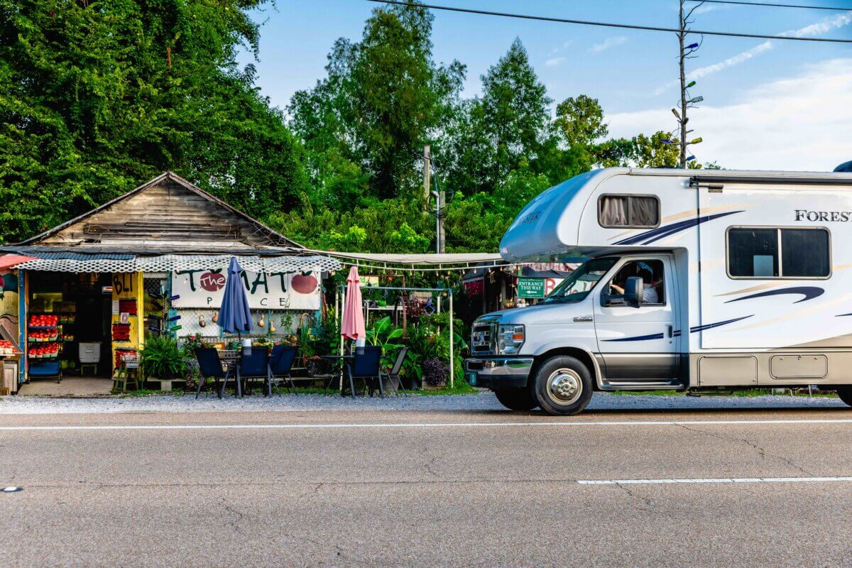 A motorhome is parked in front of a roadside produce stand called "The Tomato Place" in Vicksburg, Mississippi, with outdoor tables and umbrellas, surrounded by greenery.