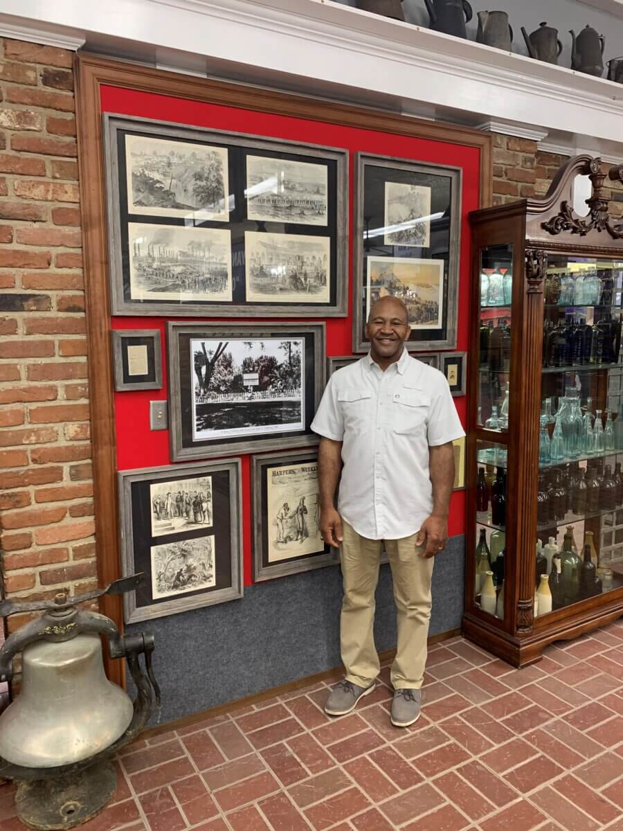 A man stands smiling next to a display of framed historical photos and prints on a red and gray wall in a Vicksburg museum, showcasing Mississippi’s rich history with brick walls and glass display cabinets—a must-see for travel enthusiasts.
