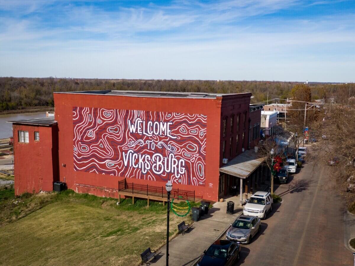 Red brick building with a large mural reading "Welcome to Vicksburg" in white lettering, located on a street with parked cars and trees, under a clear blue sky.