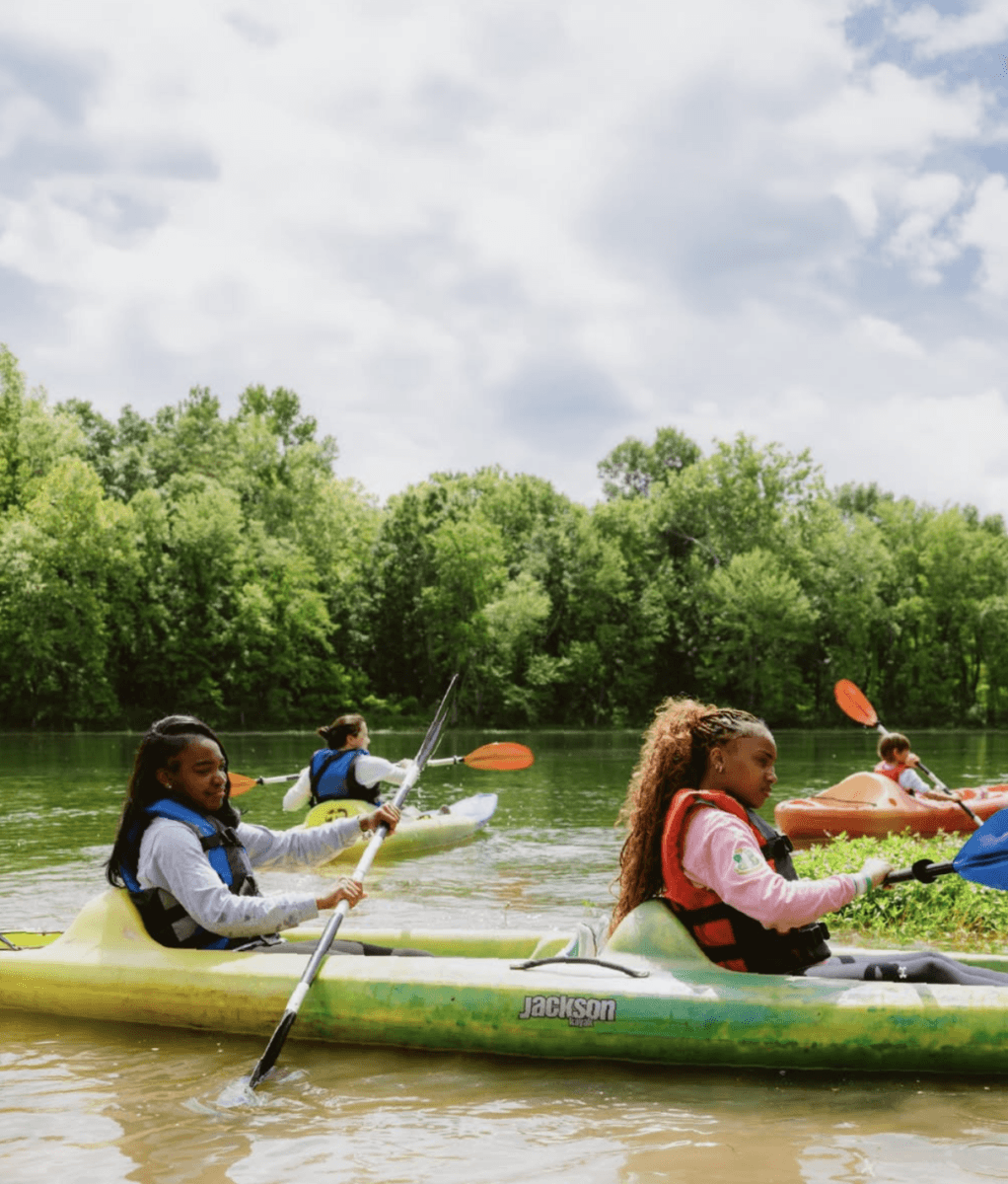 6 Ways to Get on the Water in Memphis - 2 Several people paddle kayaks on a calm river surrounded by green trees under a partly cloudy sky.