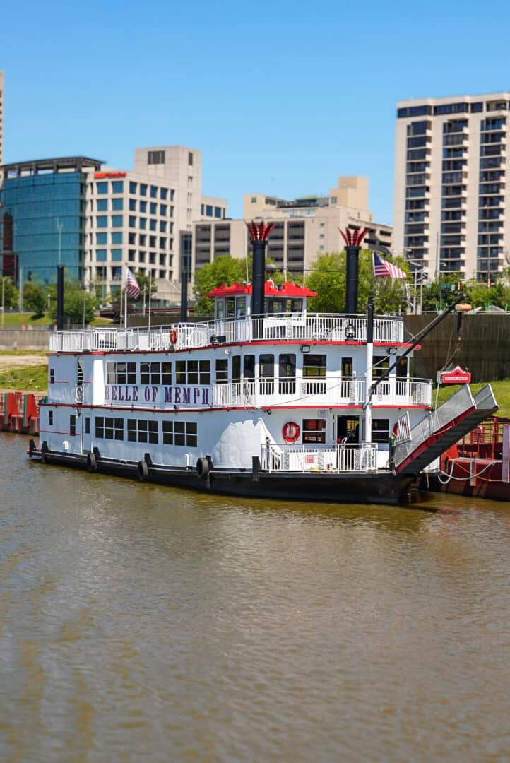 6 Ways to Get on the Water in Memphis - 5 A white riverboat named "Belle of Memphis" is docked on a river, with several flags and city buildings visible in the background.