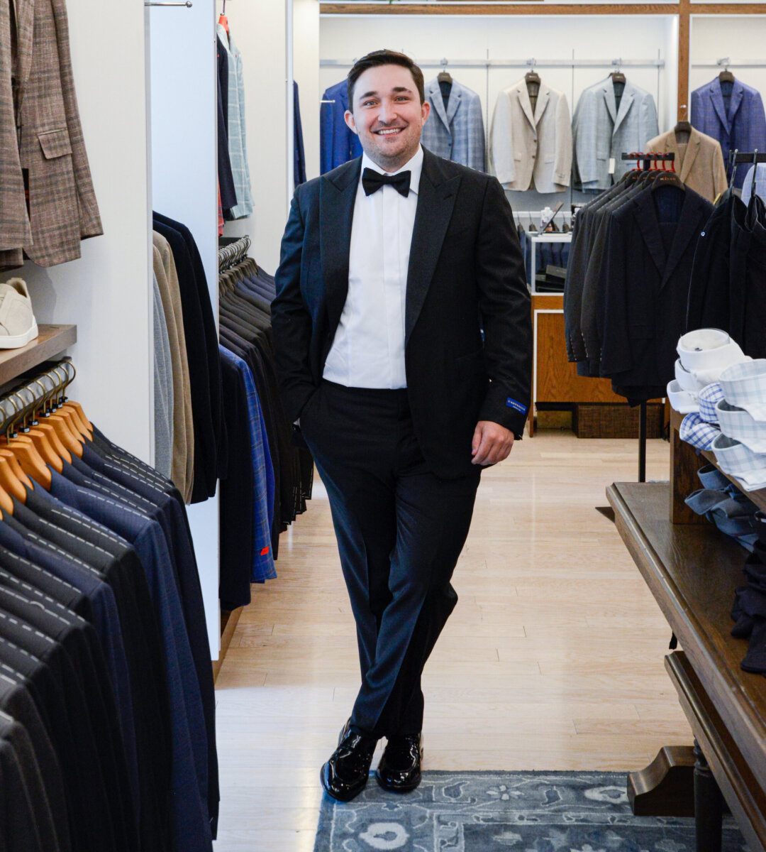 The Event-Season Trends Nashville Boutiques Are Loving Right Now - 10 All Dressed Up in a black tuxedo and bow tie, a man stands and smiles in a men's clothing store, surrounded by suits and dress shirts.