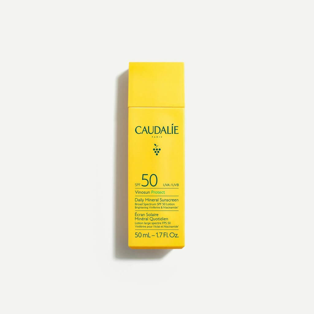 A yellow bottle of Caudalie SPF 50 Vinoperfect Daily Mineral Sunscreen, 50 mL, stands upright against a plain white background—an essential for summer beauty tips and flawless makeup for heat.