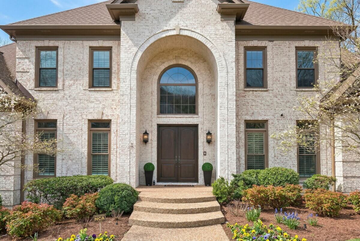 Just Listed! This Brentwood Estate Was Designed for Gathering, Entertaining & Everyday Living - 3 Front view of a Brentwood Estate brick house with double door entrance, arched window above, symmetrical windows, landscaped shrubs, and a stone walkway—perfect for entertaining or everyday living.