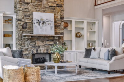 A Brentwood Estate-inspired living room with a stone fireplace, abstract art, two light-colored sofas, built-in shelves, a round coffee table, and woven baskets—perfect for everyday living and entertaining.