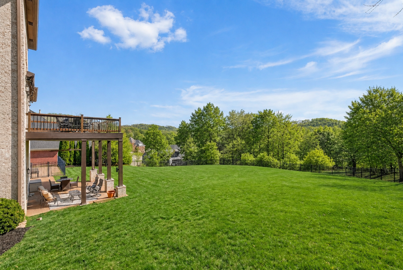 Just Listed! This Brentwood Estate Was Designed for Gathering, Entertaining & Everyday Living - 20 Spacious backyard at 513 Arden Wood Place with green lawn, patio seating area under a wooden deck, and trees in the background beneath a blue sky dotted with clouds.