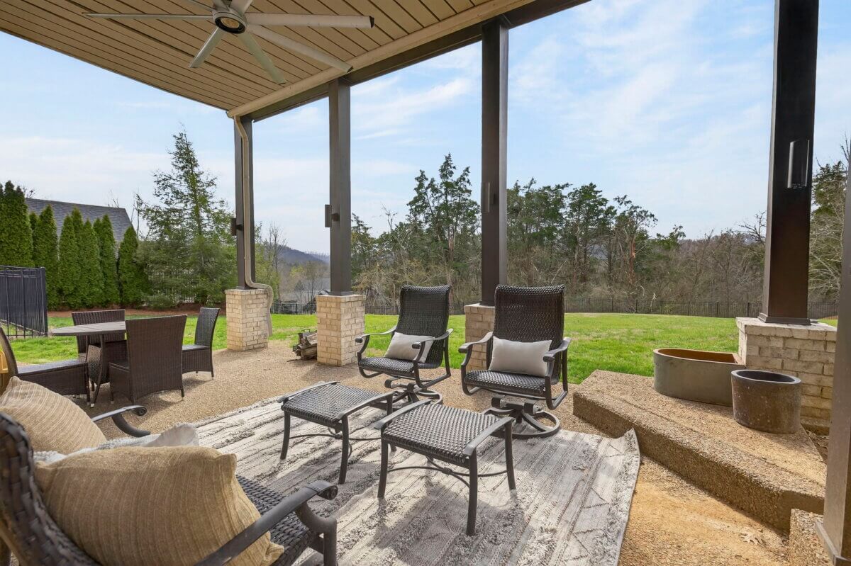 Just Listed! This Brentwood Estate Was Designed for Gathering, Entertaining & Everyday Living - 19 Covered patio with a ceiling fan, wicker chairs, small tables, and outdoor rug—perfect for everyday living or entertaining—overlooking a grassy backyard with trees in the background at Brentwood Estate.