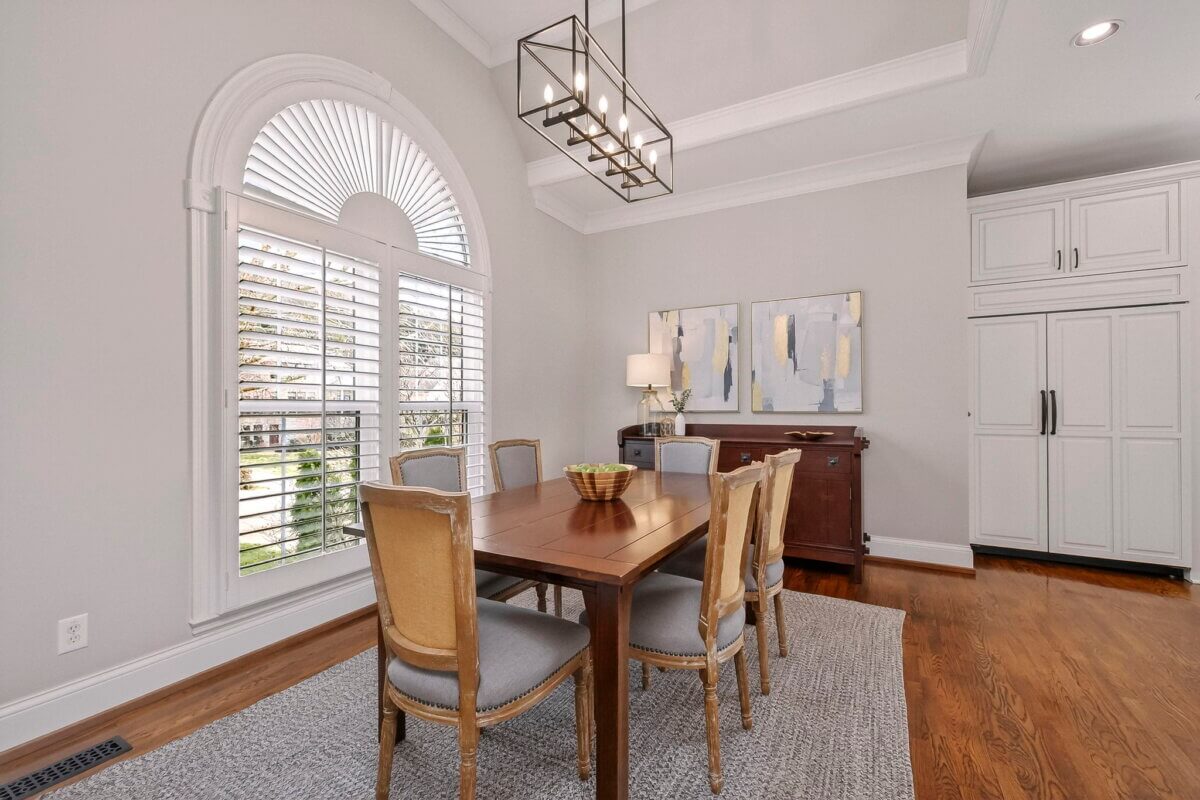 Just Listed! This Brentwood Estate Was Designed for Gathering, Entertaining & Everyday Living - 11 A dining room designed for everyday living and entertaining, featuring a wooden table, six chairs, a gray rug, arched window with shutters, chandelier, wooden sideboard, and modern wall art.