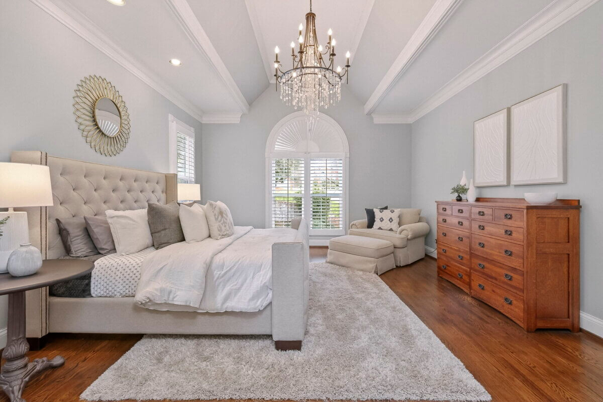 Just Listed! This Brentwood Estate Was Designed for Gathering, Entertaining & Everyday Living - 12 A spacious, elegant bedroom in Brentwood Estate features a tufted bed, chandelier, large window, wood dresser, armchair, and neutral decor—perfect for everyday living.
