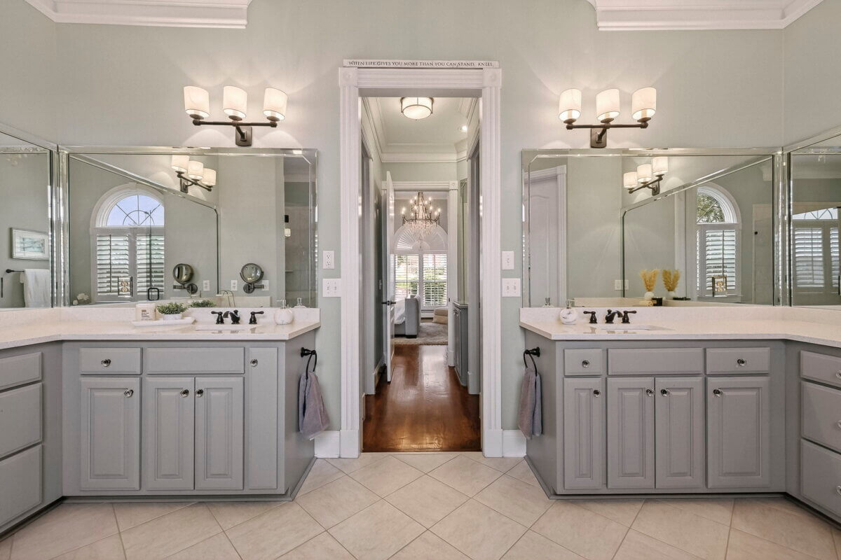 Just Listed! This Brentwood Estate Was Designed for Gathering, Entertaining & Everyday Living - 14 Double vanity bathroom with gray cabinets, large mirrors, wall-mounted lights, and a doorway leading to a bedroom with a chandelier and plantation shutters—perfect for everyday living in a Brentwood Estate.