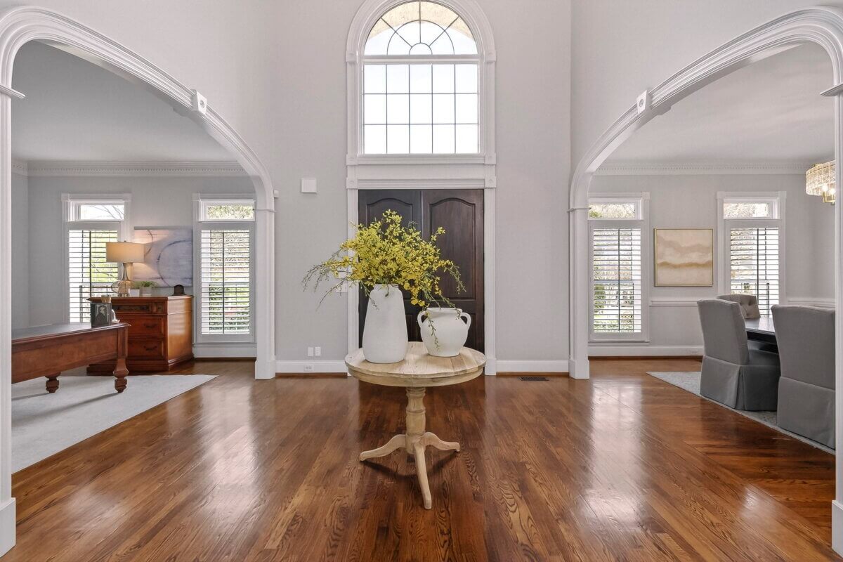 Just Listed! This Brentwood Estate Was Designed for Gathering, Entertaining & Everyday Living - 4 Spacious foyer with wood floors, perfect for entertaining, features a round table holding white vases with yellow flowers and large arched windows letting in natural light.